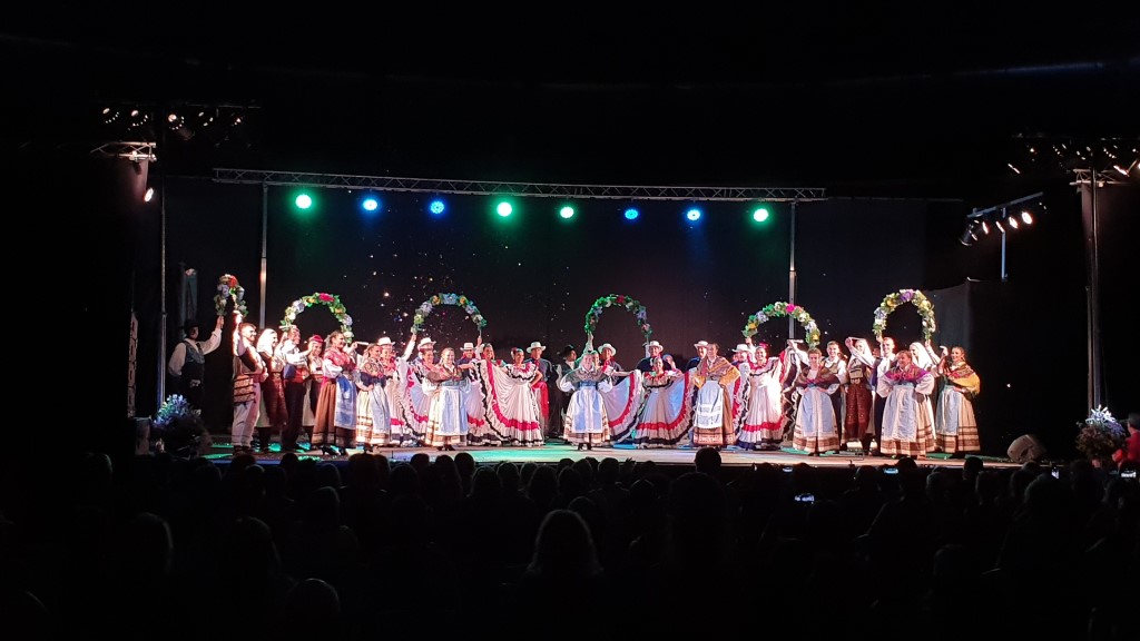 Foto de una representación de danzas en el Festival Internacional de Arroyo de la Encomienda. Sobre el escenario se puede ver a un conjunto de danzas de hombres y mujeres. Algunos de los miembros sujetan seis arcos de flores y bajo cada uno de ellos danza una bailarina.